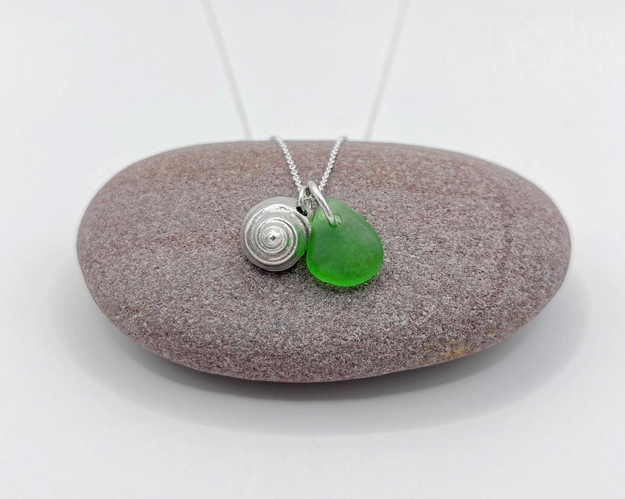 Necklace with a silver shell and green sea glass laid on a purple pebble against a white background