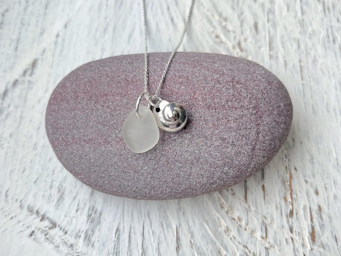 Necklace with a silver shell and white sea glass pendant laid on a purple pebble against a textured surface
