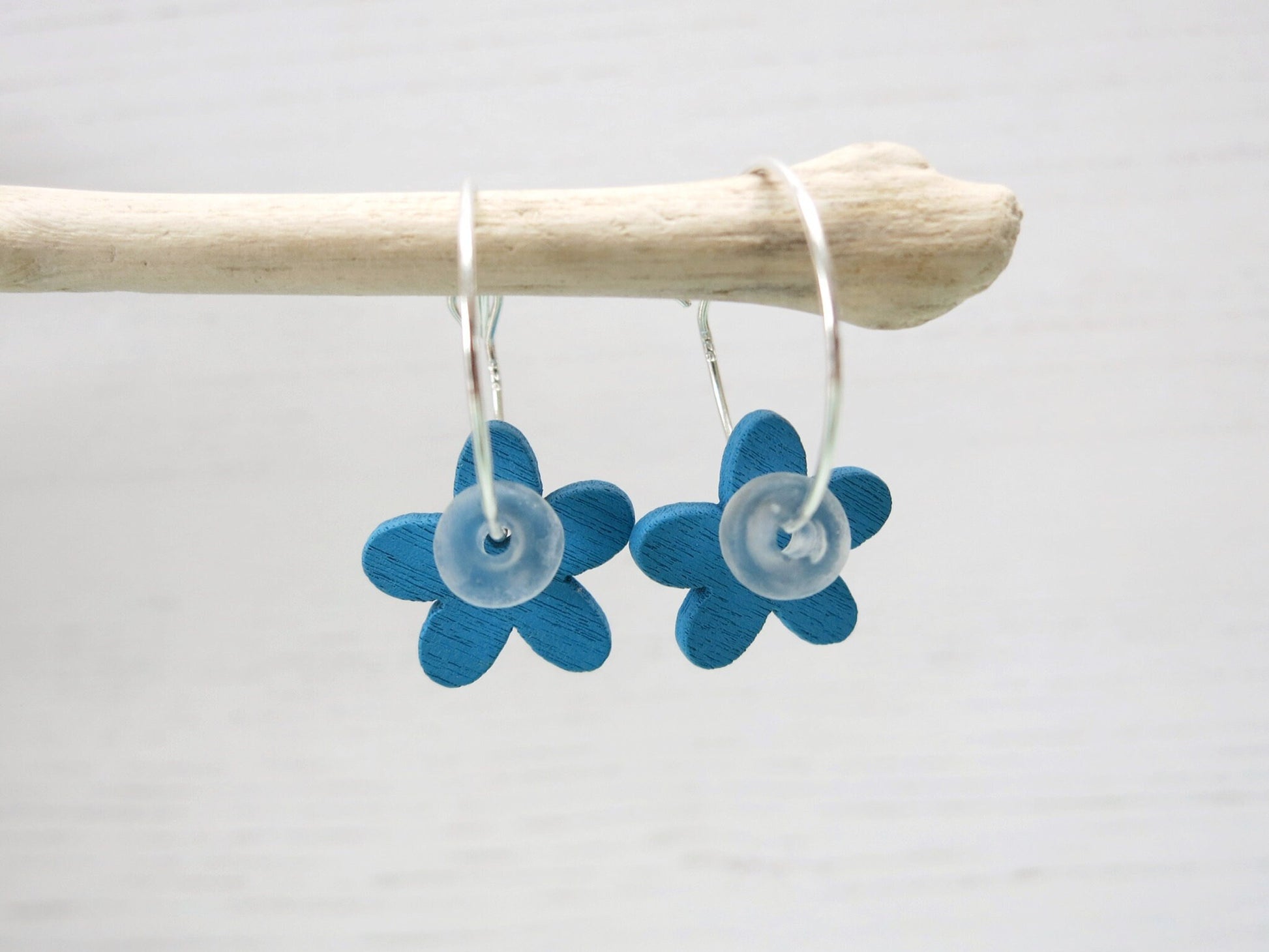 Silver hoop earrings with Blue wooden flowers and a glass bead hanging on driftwood against a white background