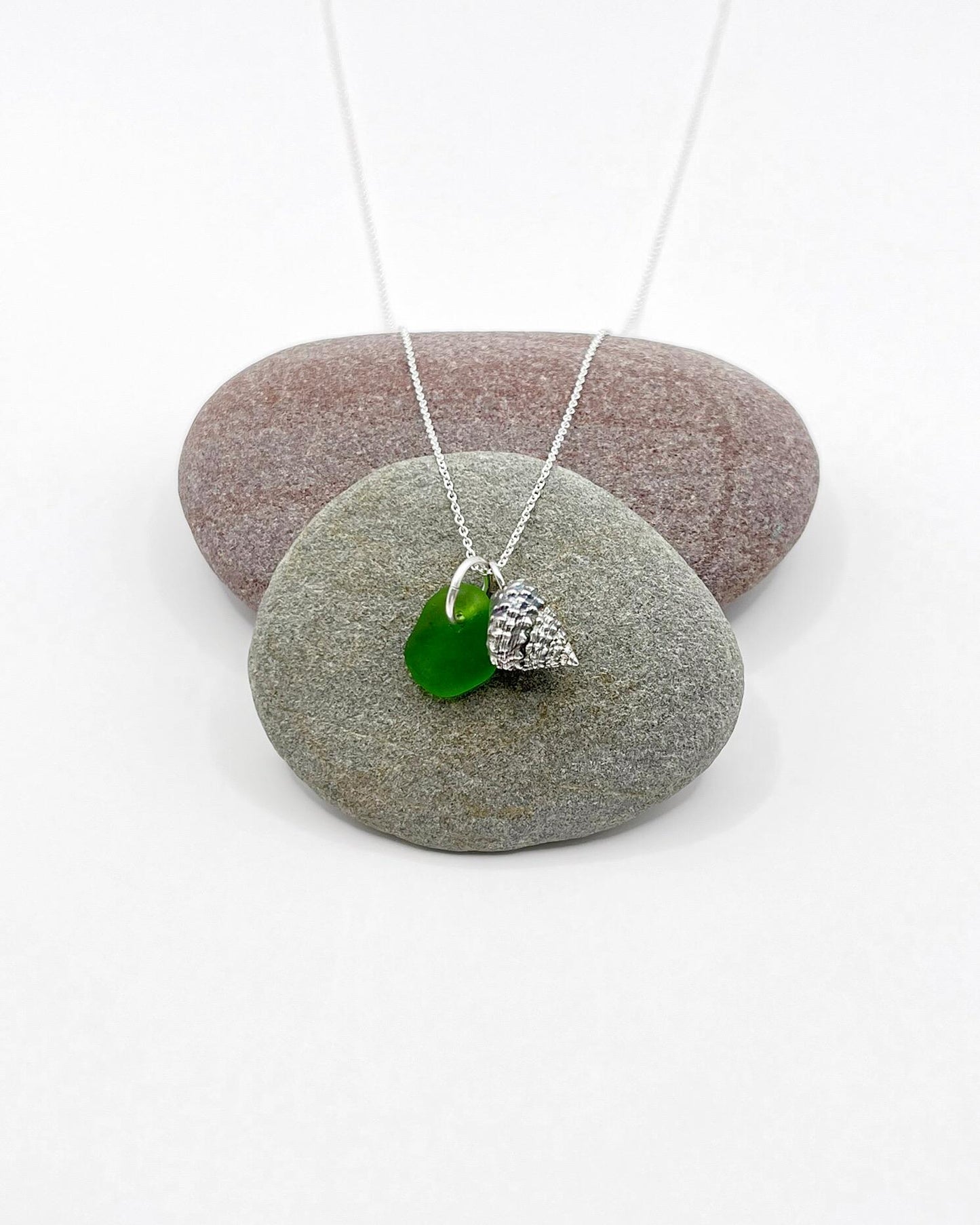 Necklace with green sea glass and silver shell pendant laid across two pebbles against a white background
