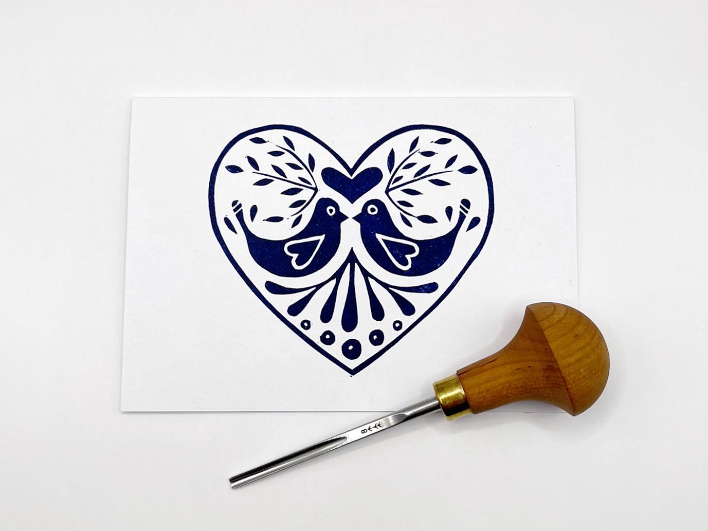 White card with an original lino printed blue heart design featuring two birds laid on a white background. Photo includes lino cutting tool.