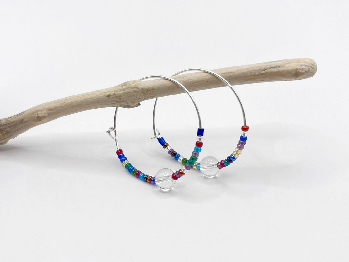 Colourful beaded hoop earrings on a white background hanging on a piece of driftwood.