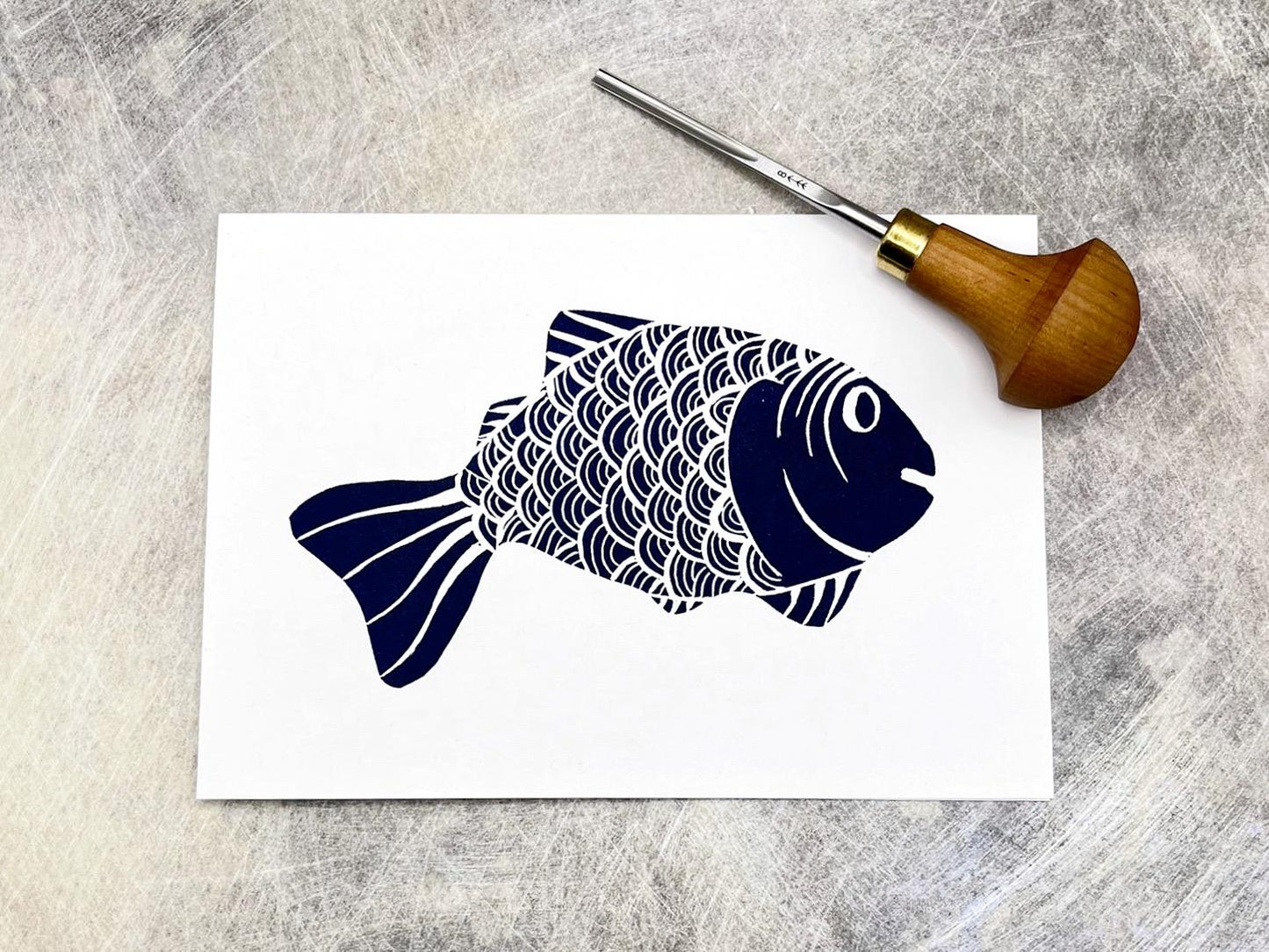 White card with an original lino printed blue fish, laid on a textured background. Photo includes the lino cutting tool used.