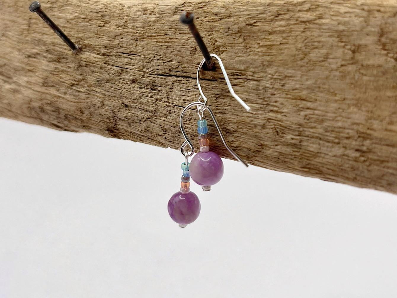 Pair of pastel seed bead and lilac jasper earrings hanging on driftwood against a white background.