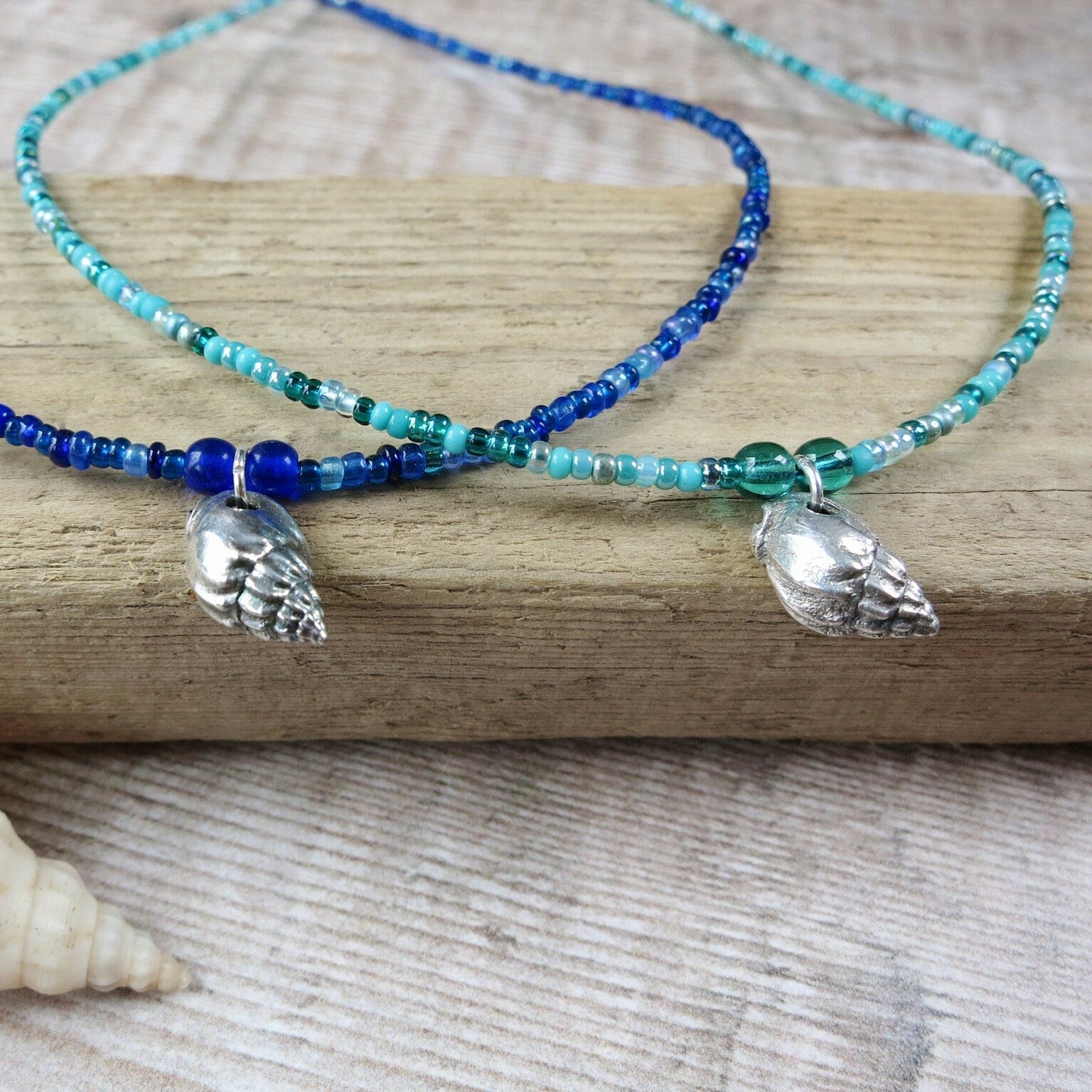 Two seed bead necklaces with shell pendants laid on driftwood with a shell in the foreground.