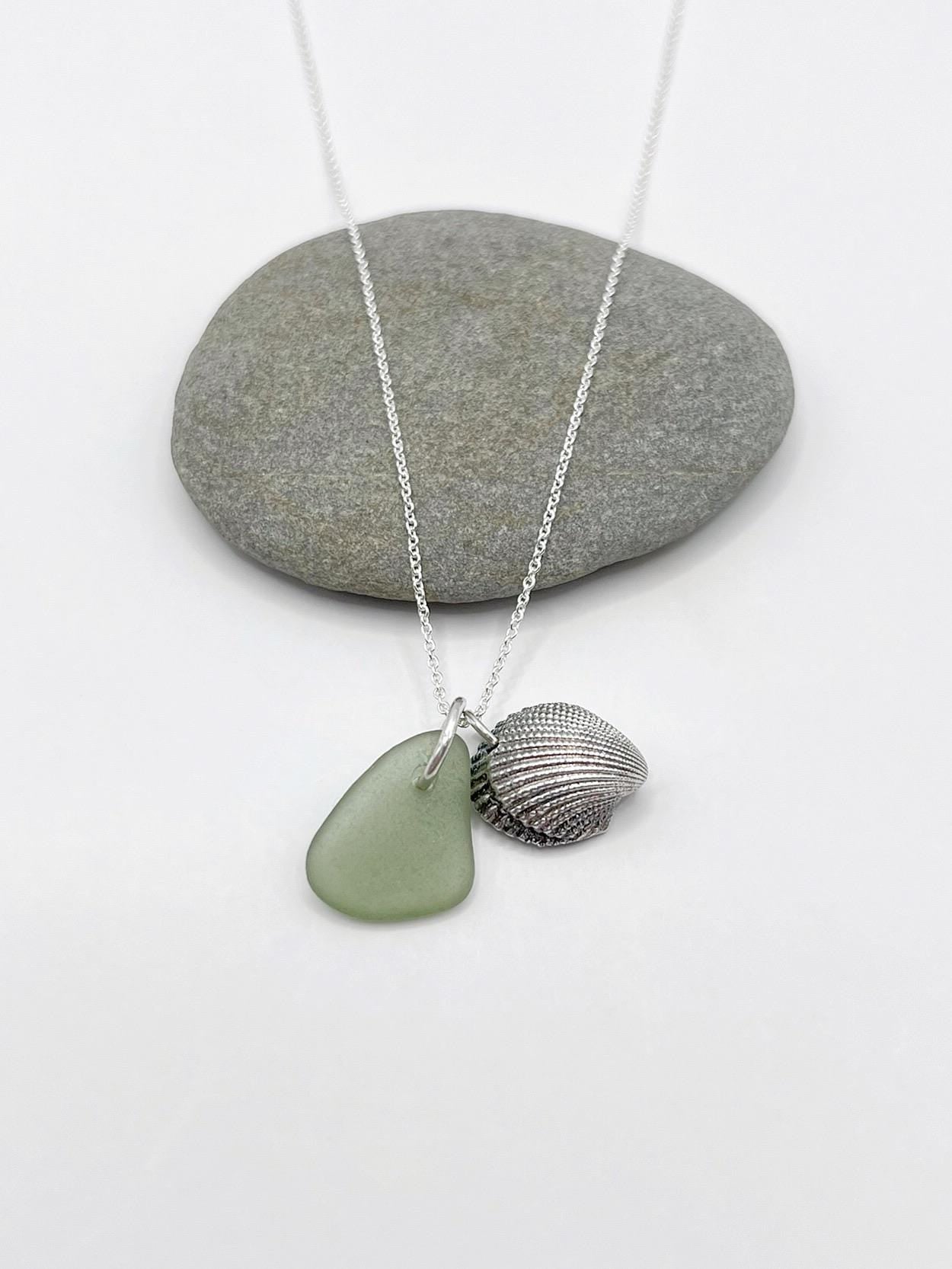 Necklace with green sea glass and a silver shell charm laid on a pebble with a white background