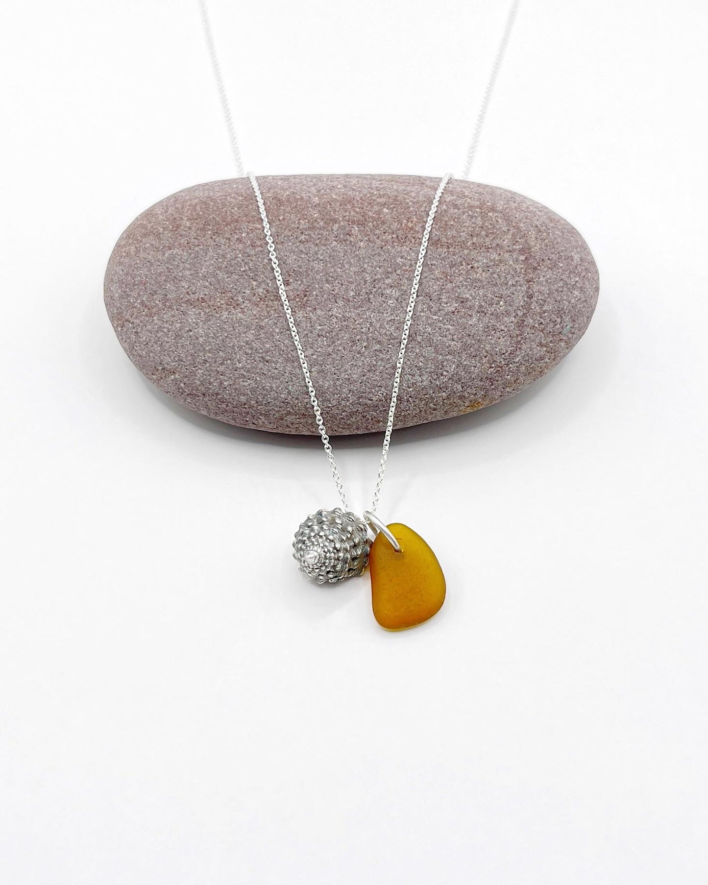 Necklace with burnt orange sea glass and silver shell pendant laid in front of a purple pebble against a white background