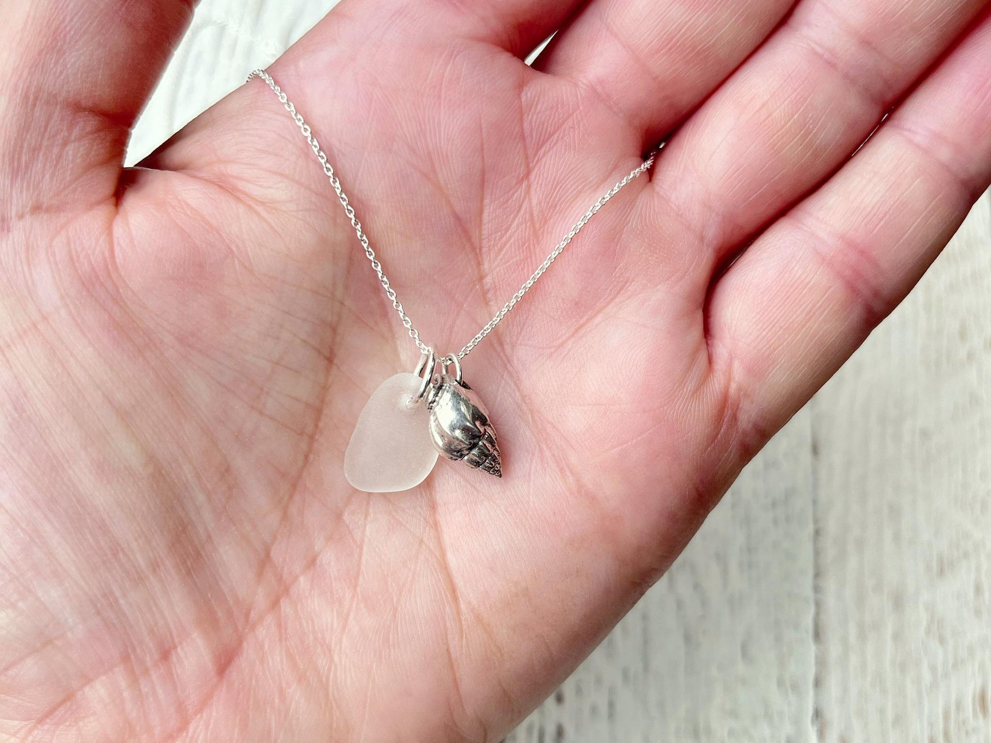 Hand holding a necklace with white sea glass and silver shell pendant against a textured surface