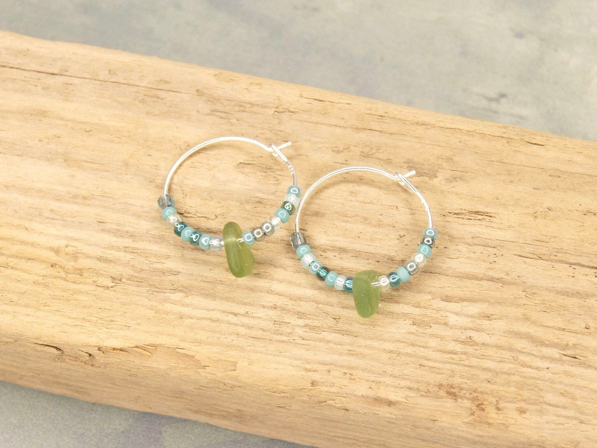 Turquoise seed bead hoop earrings with green sea glass laid on a driftwood.