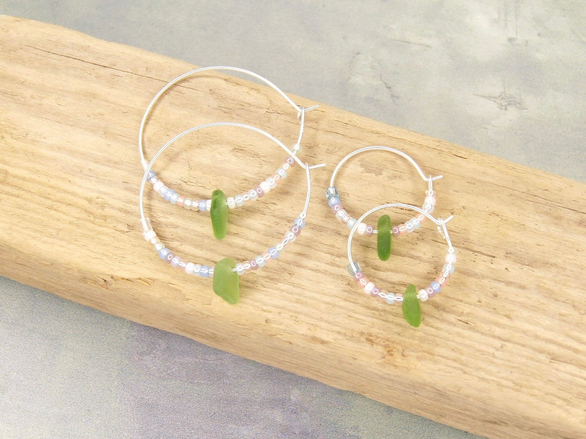 Two pairs of hoop earrings with pastel seed beads and green beads laid on driftwood.