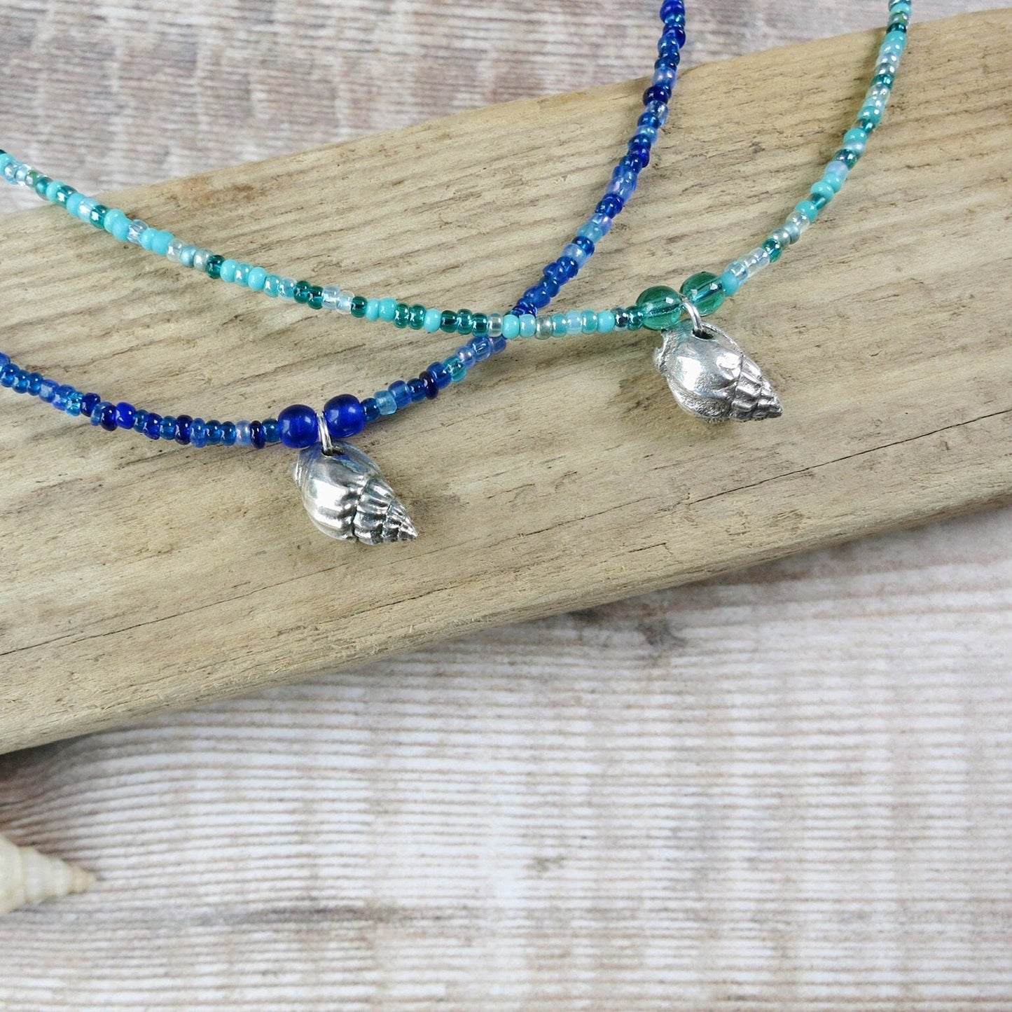 Two seed bead necklaces with silver shell pendants laid on driftwood against a textured surface