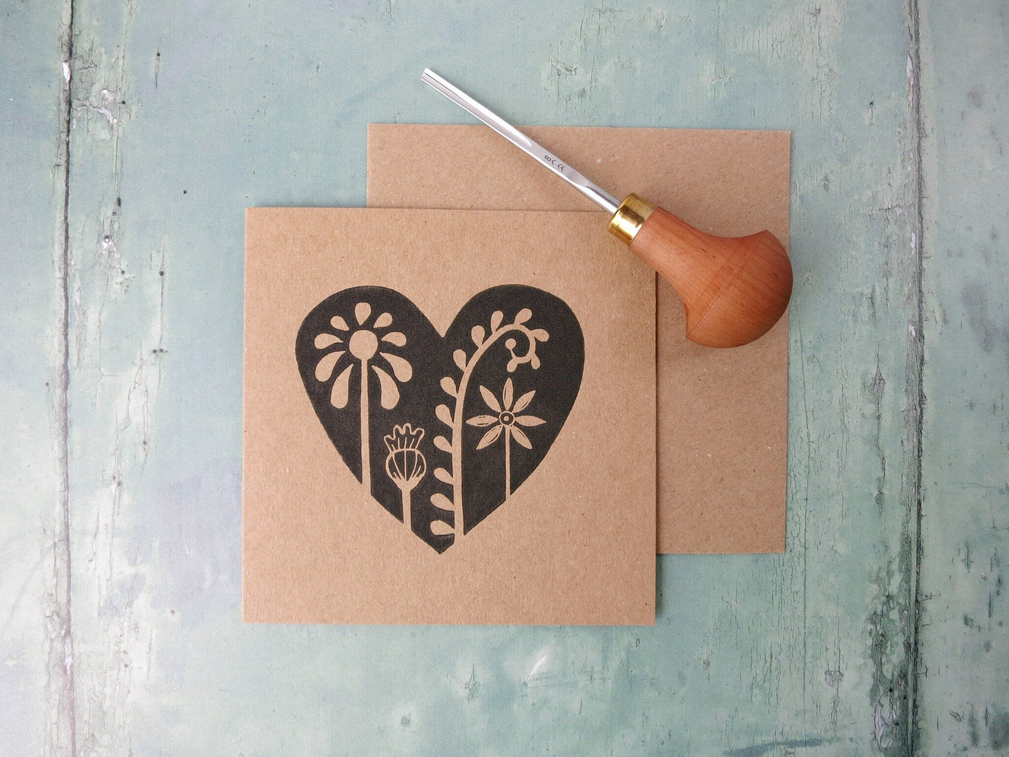Kraft paper card with an original lino printed floral heart design laid on  a textured surface. Photo includes the lino cutting tool used.