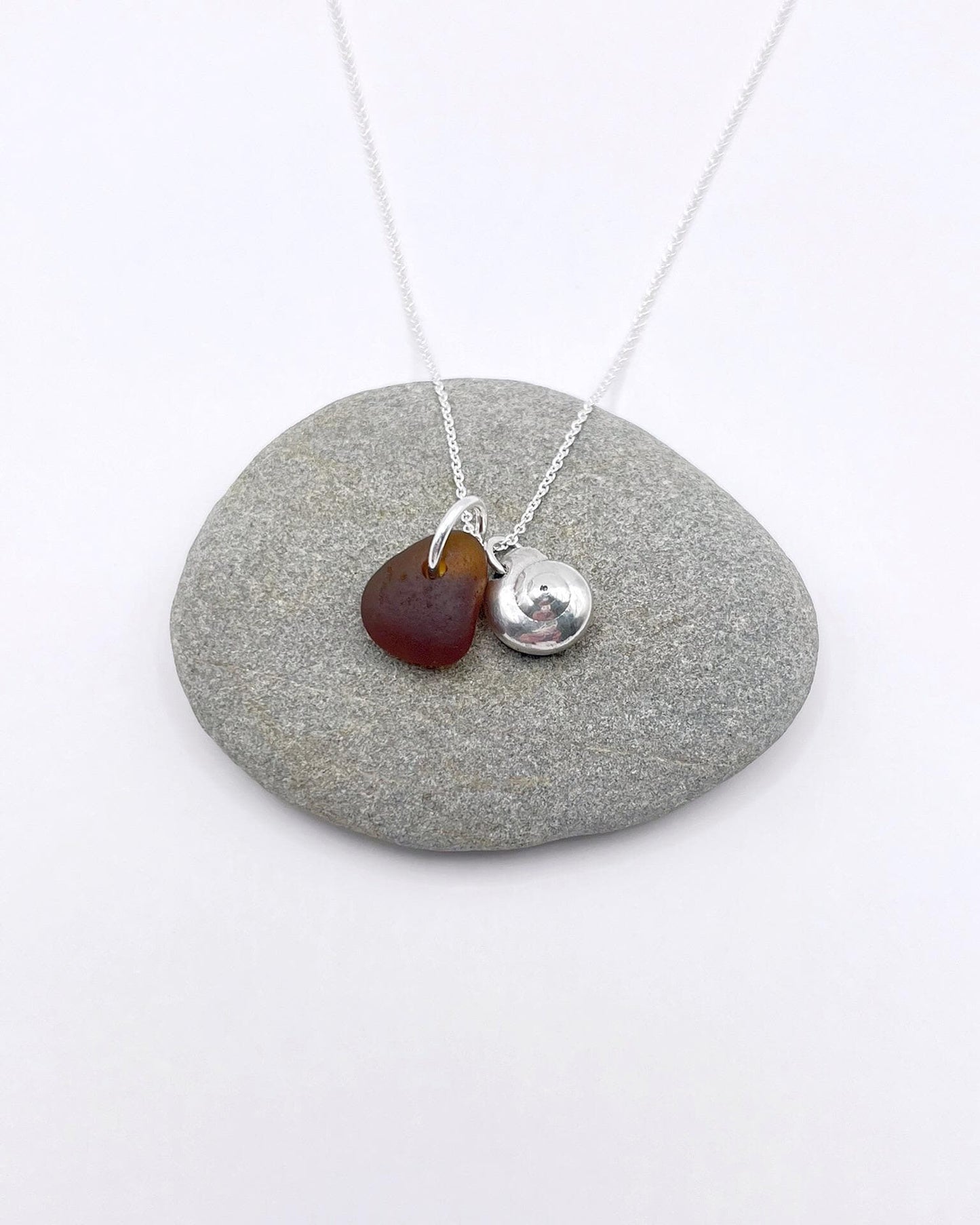 Necklace with burnt orange sea glass and a silver shell pendant, laid on a grey pebble on a white surface