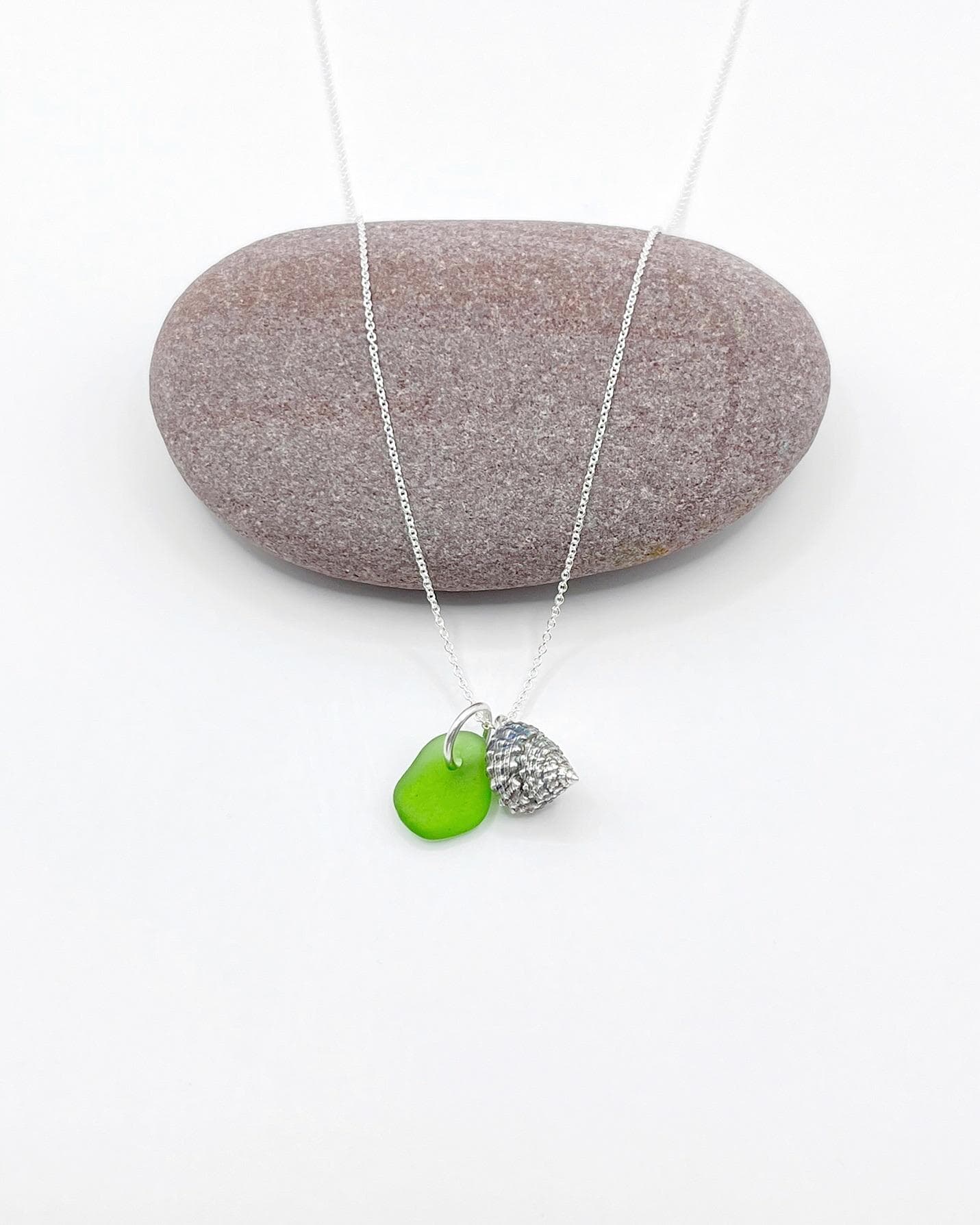 Necklace with green sea glass and silver shell pendant laid in front of a purple pebble on a white background