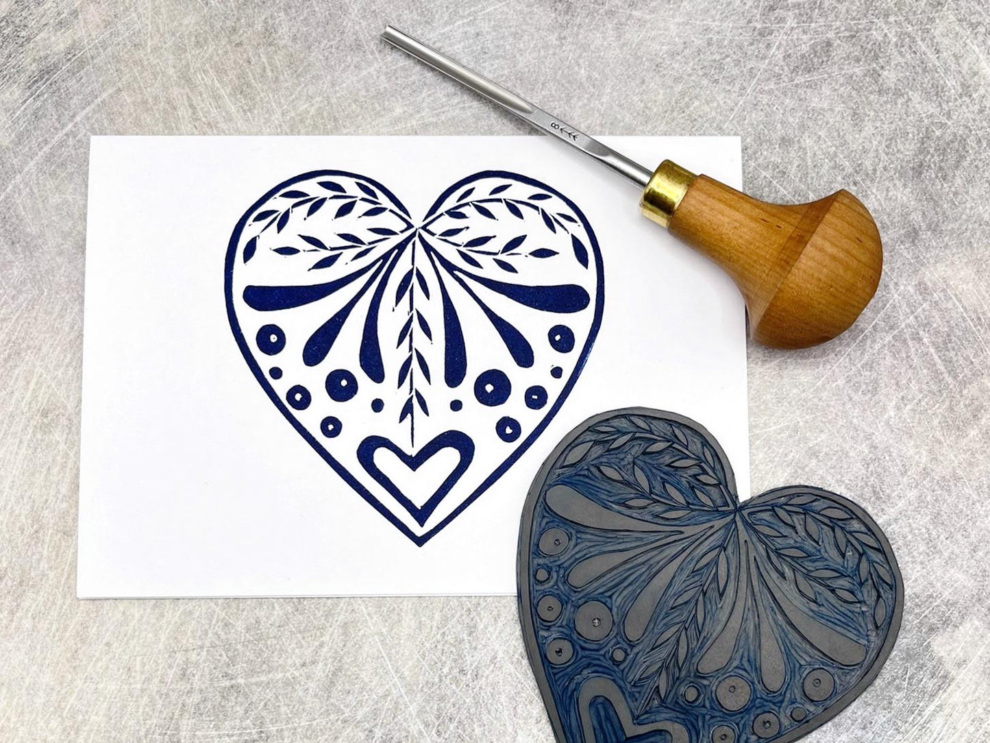 White card with an original lino printed  blue heart design laid on a textured surface. Photo includes lino and cutting tool.