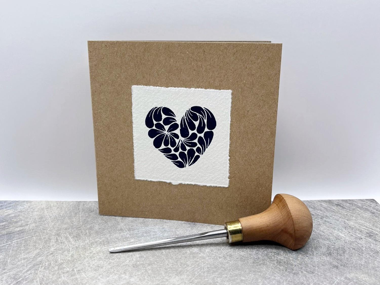 Kraft card with original lino printed blue heart design on white paper stood against a white background. Photo includes lino cutting tool.
