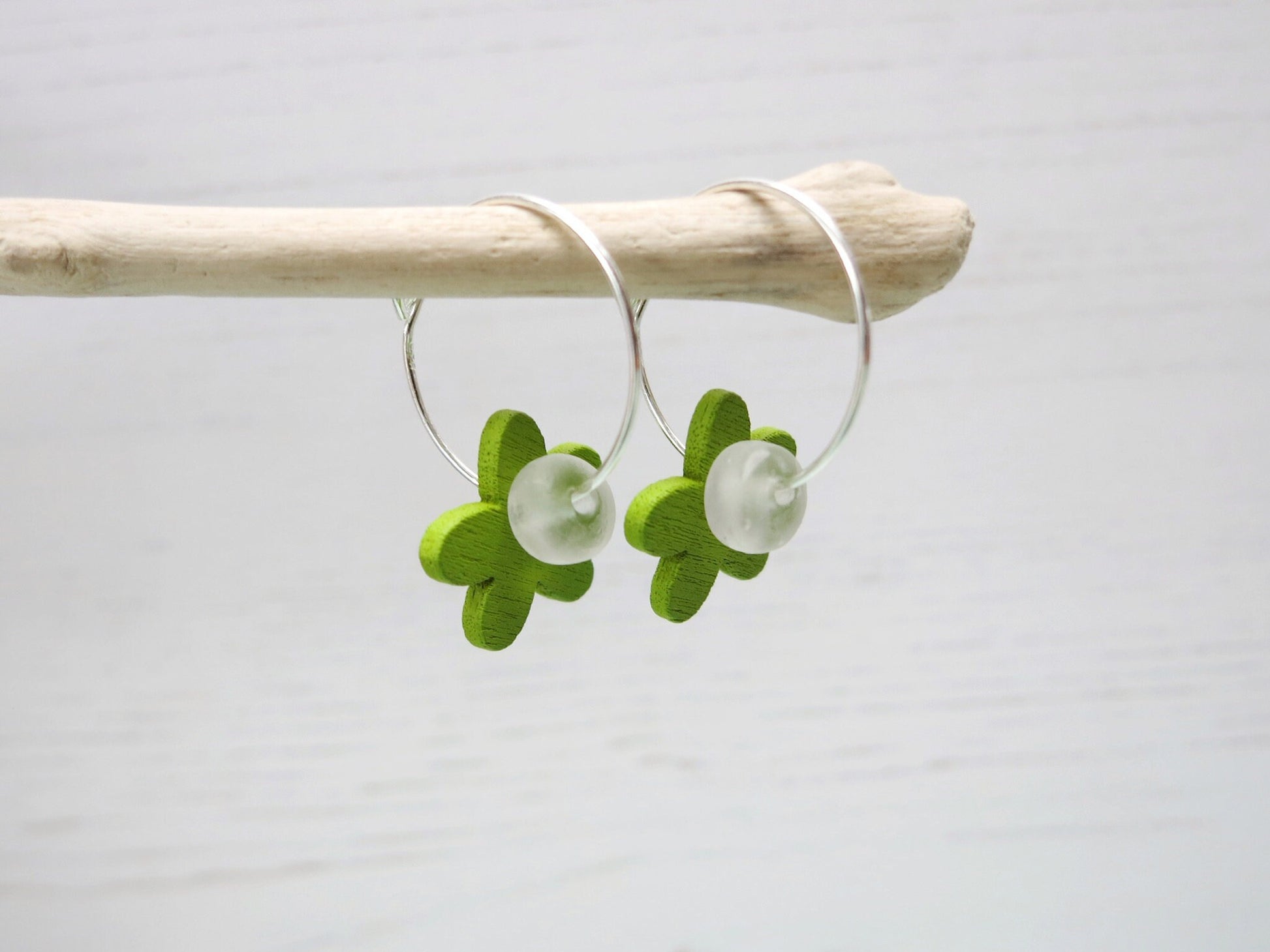 Side view of silver hoop earrings with green wooden flowers and a glass bead hanging on driftwood against a white background