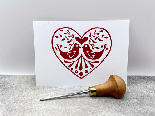 White card with red original lino printed heart design featuring two birds. Photo includes lino cutting tool used stood against a textured background.
