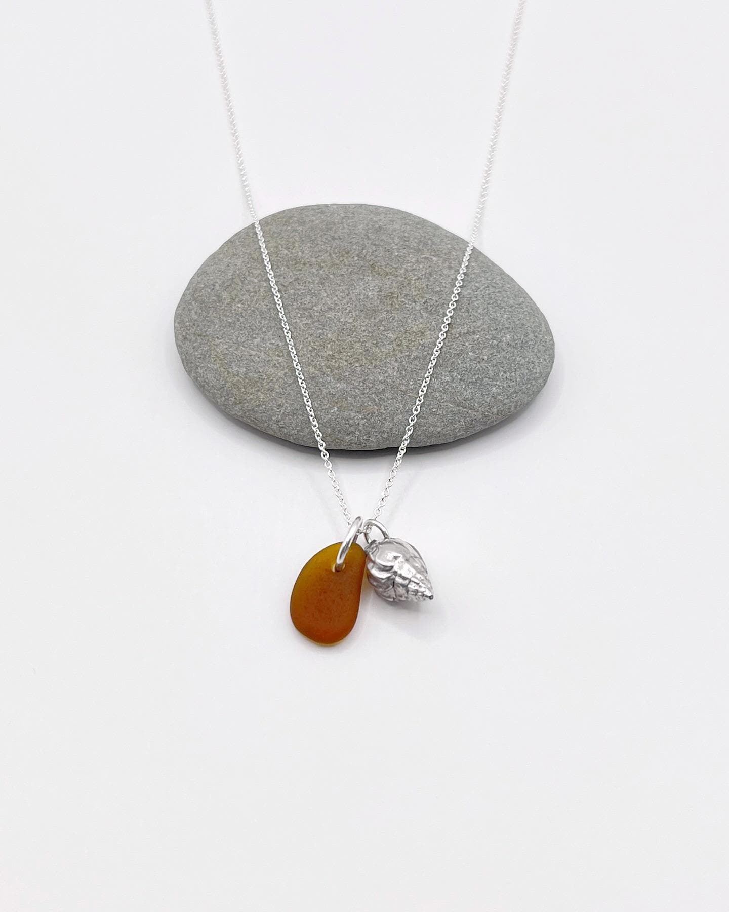 Necklace with burnt orange sea glass and silver shell pendant laid in front of a grey pebble on a white background