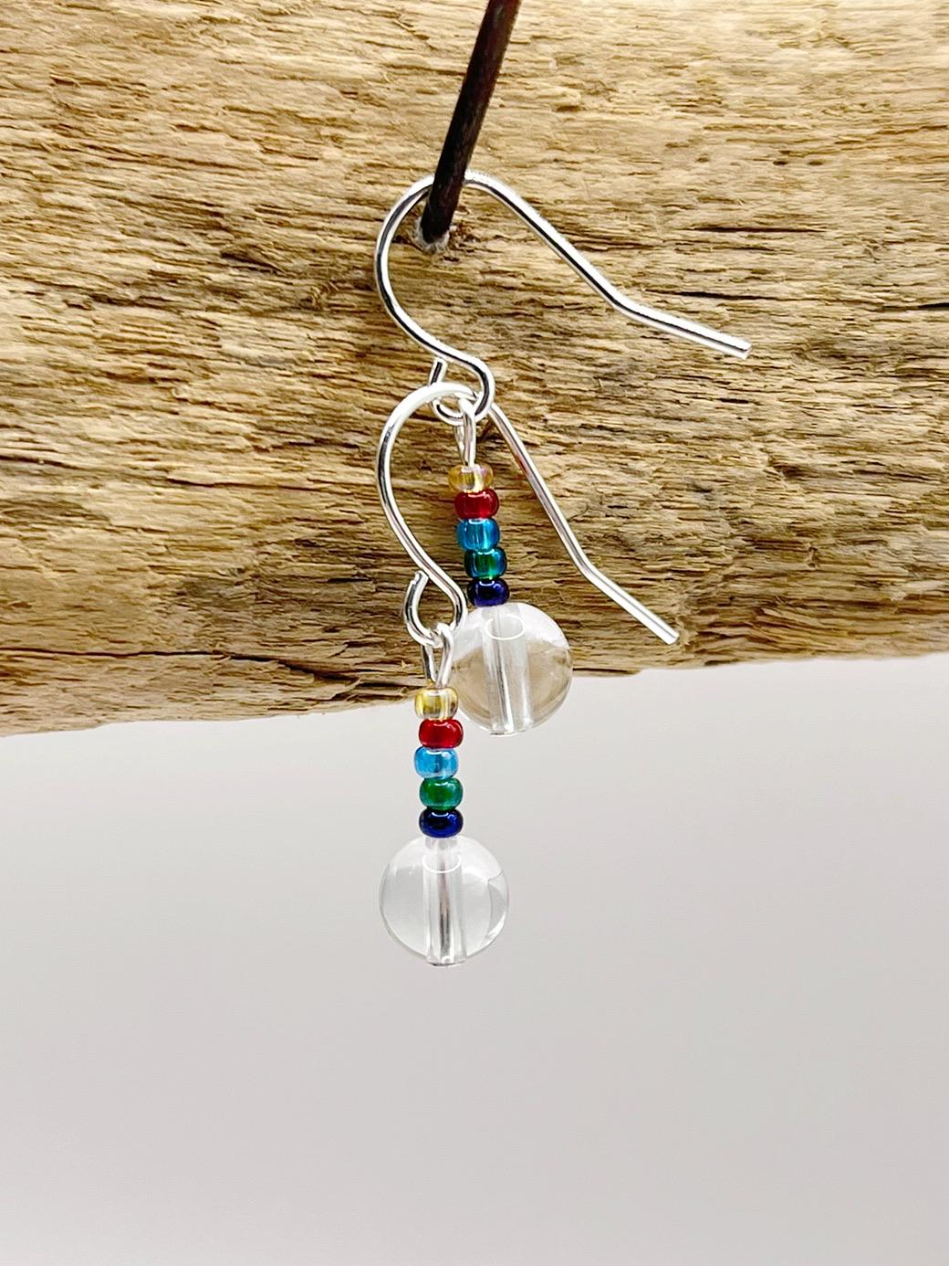 Silver earrings with clear crystal and rainbow seed beads hanging on driftwood