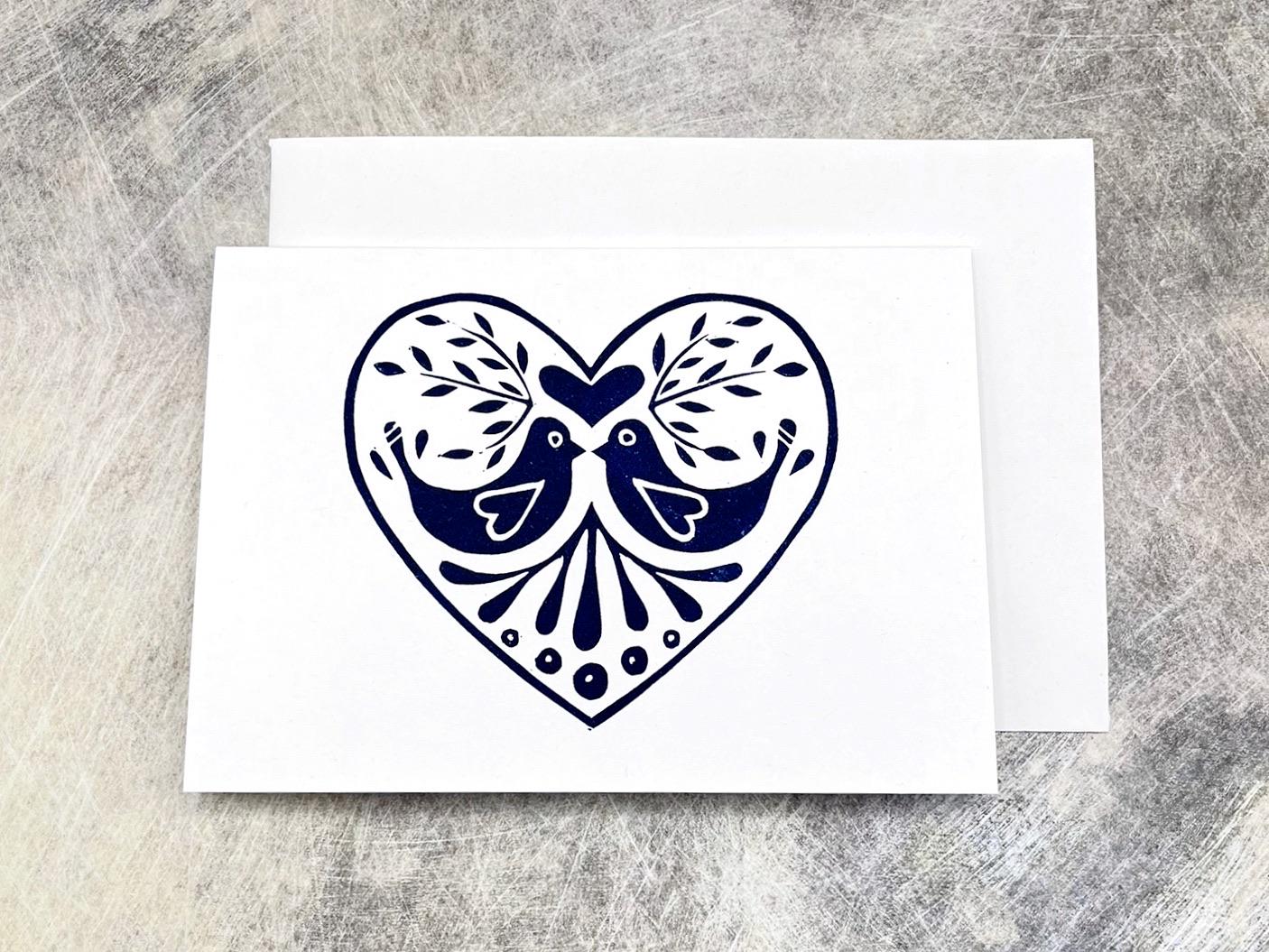 White card with an original lino printed blue heart design featuring two birds, laid on a grey textured background.