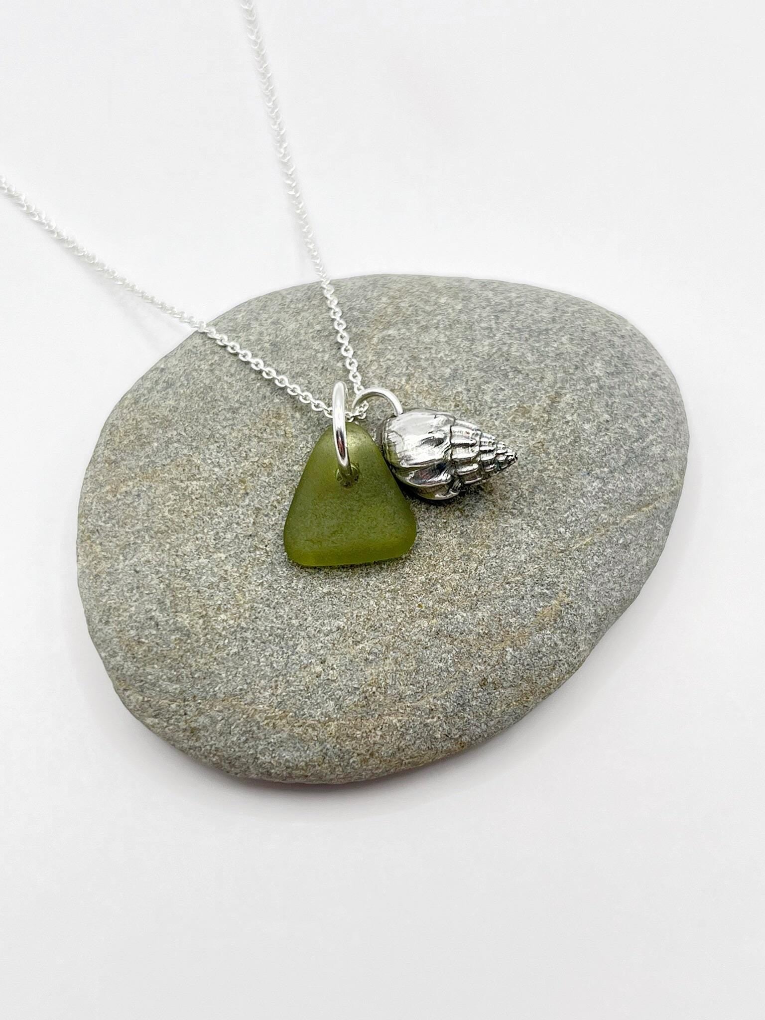 Necklace with olive green sea glass and a silver shell charm laid on a grey stone.