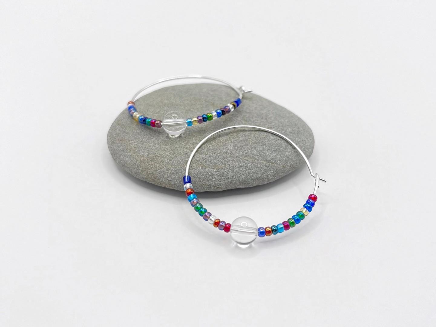 Close up of rainbow seed bead and crystal hoop earrings laid on a grey pebble against a white background.