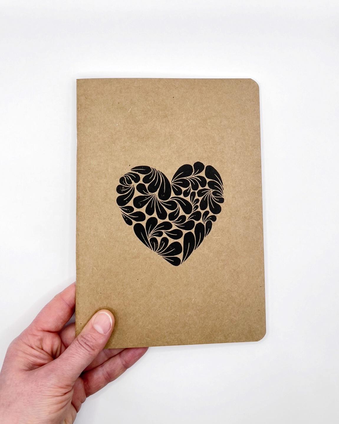 Kraft paper notebook with an original black lino printed heart design held by a hand on a white background