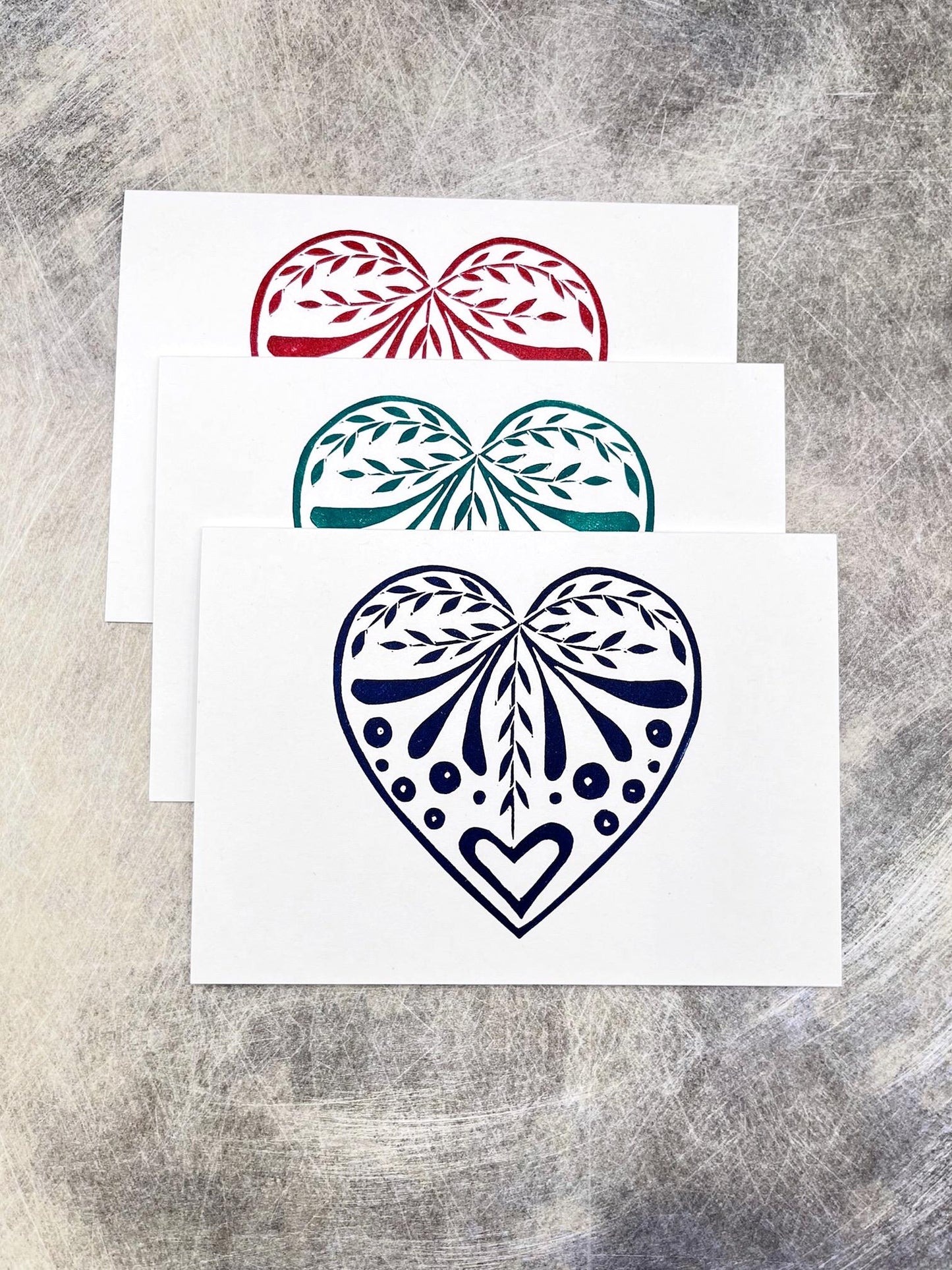 Set of three white cards with original lino printed heart designs in blue, green and red. Laid on a textured surface