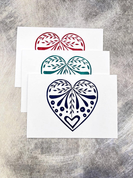 Set of three white cards with original lino printed heart designs in blue, green and red. Laid on a textured surface
