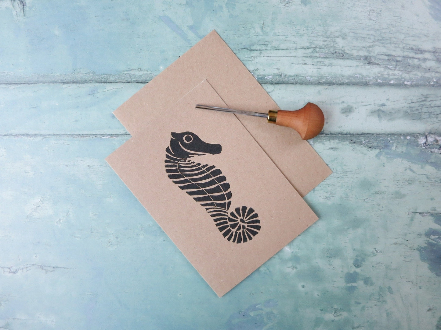 Kraft paper card with an original seahorse lino print laid at an angle on a textured blue surface. Photo includes lino cutting tool
