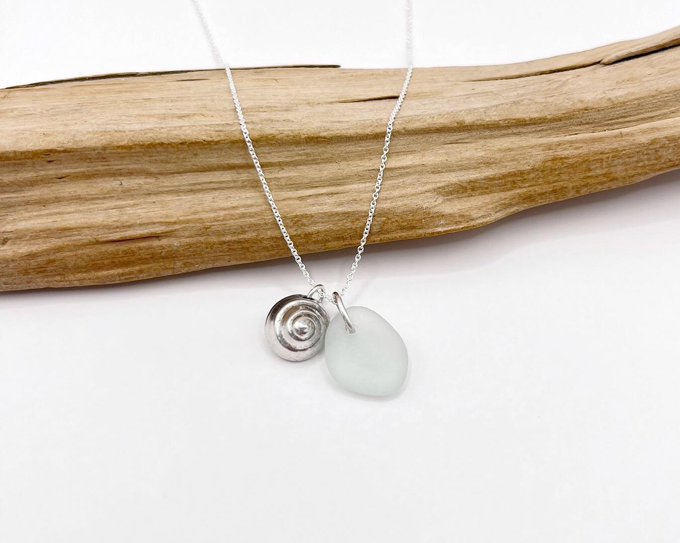 Necklace with a silver shell and seaglass pendant laid across driftwood against a white background