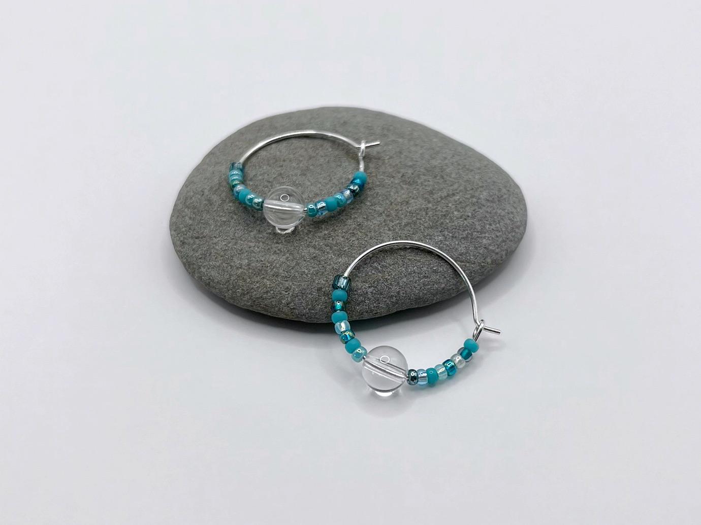 Silver hoop earrings with turquoise seed beads and crystal laid on a grey pebble against a white background