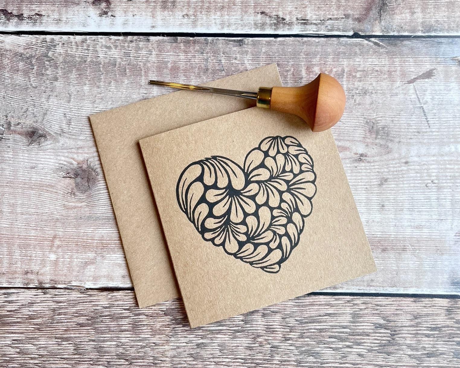 Kraft paper card with an original lino printed heart laid on a wooden surface. Photo includes lino cutting tool used.