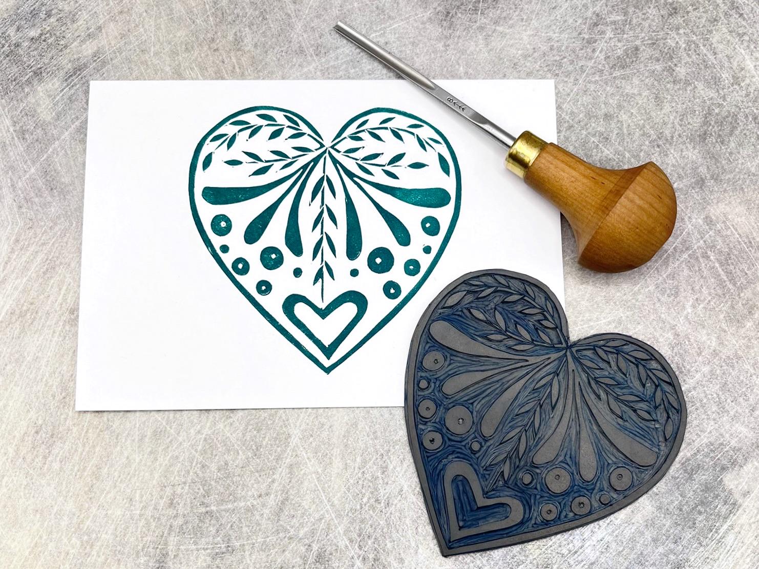 Green heart card from the set of three white cards with original lino printed heart designs. Laid on a textured surface. Photo includes lino and cutting tool.