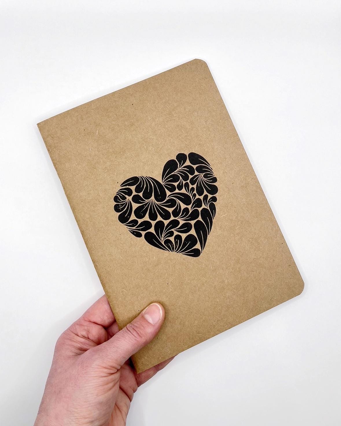 Kraft paper notebook with an original black lino printed heart design held by a hand on a white background