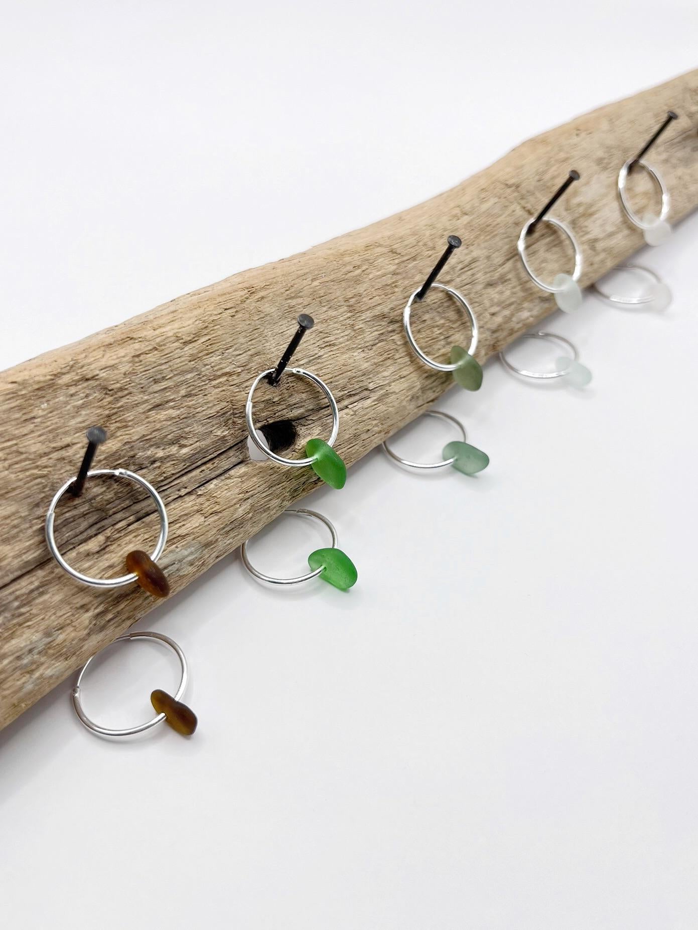 Silver hoop earrings with sea glass hanging on driftwood against a white background