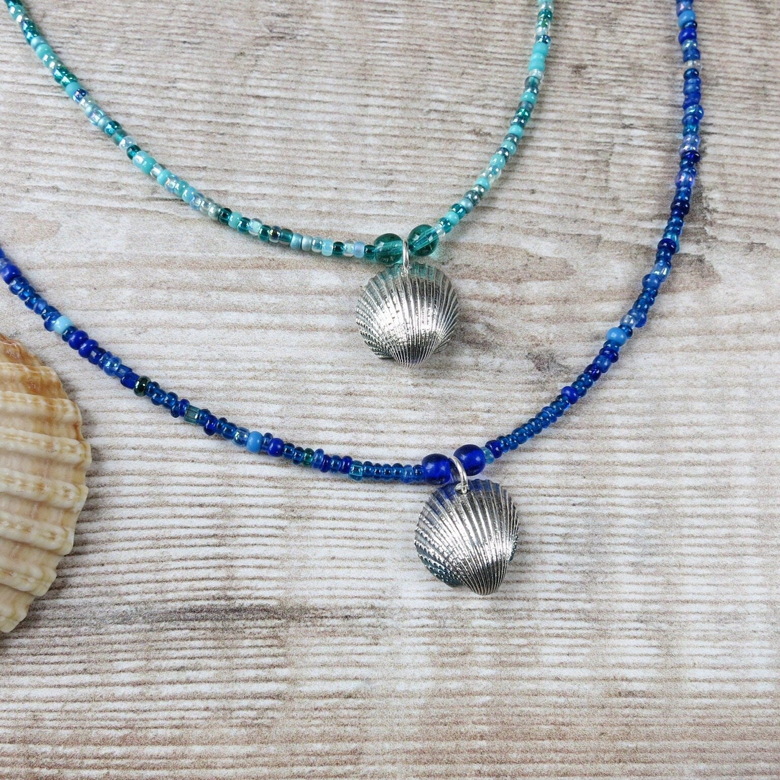 Two seed bead necklaces with silver coated shell pendants laid on a textured surface