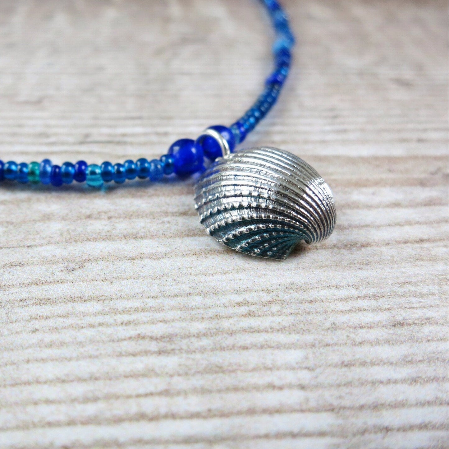 Close up of blue seed bead necklace with a silver shell pendant laid on a textured beige surface