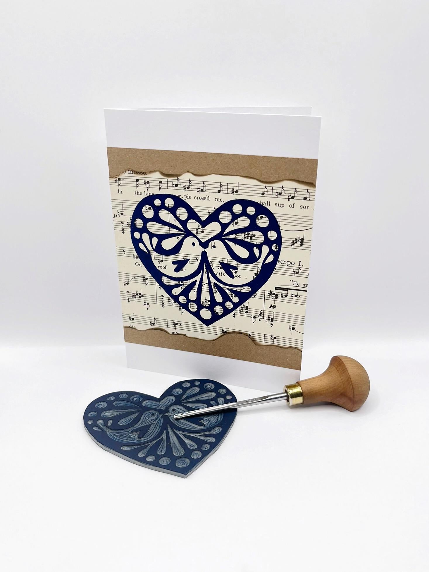 Card with original lino printed heart design on sheet music. Photo includes lino and cutting tool used against a white background.