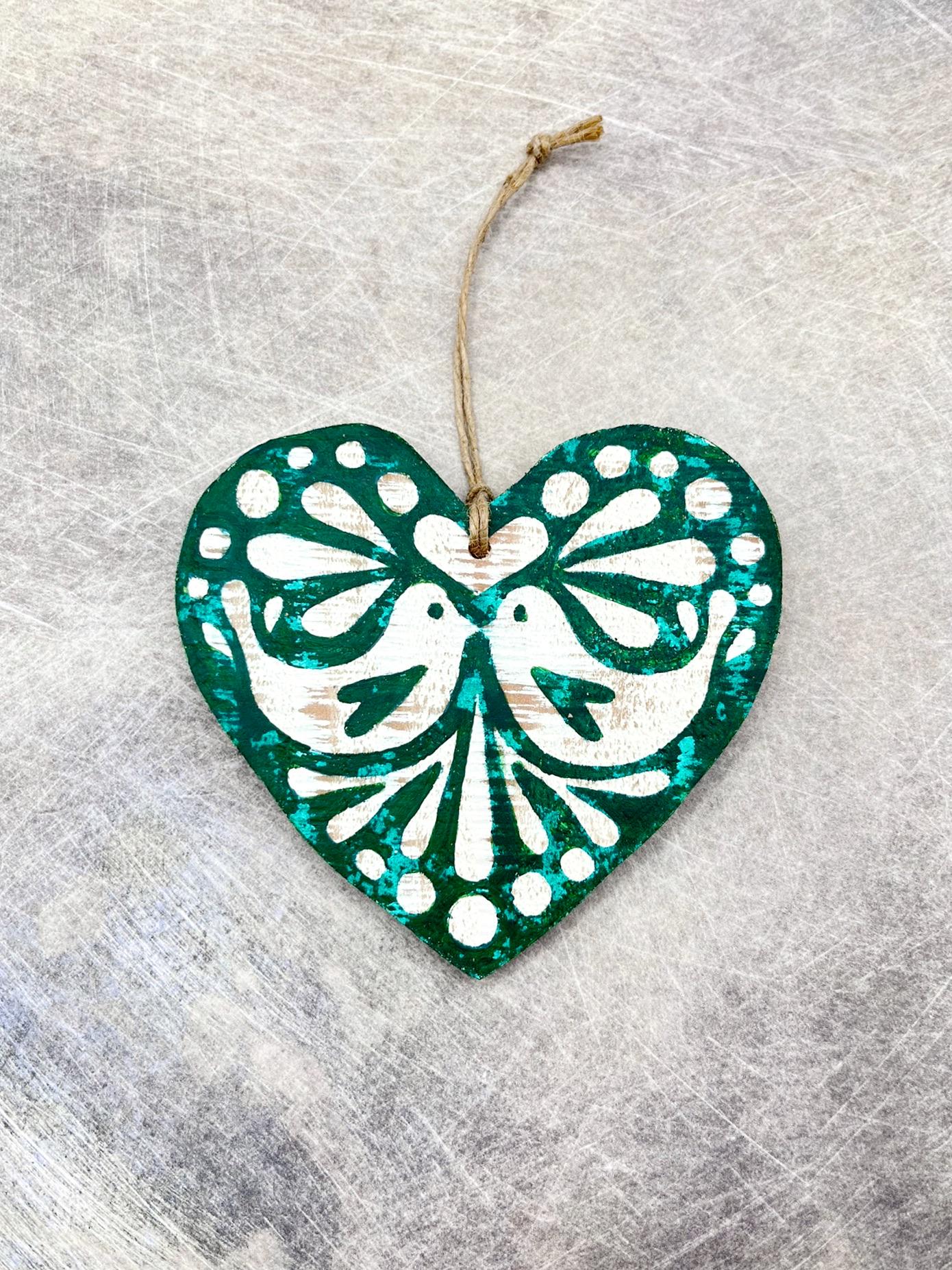 Green and white heart-shaped wooden hanging decoration with folk style love bird print laid on a textured surface.