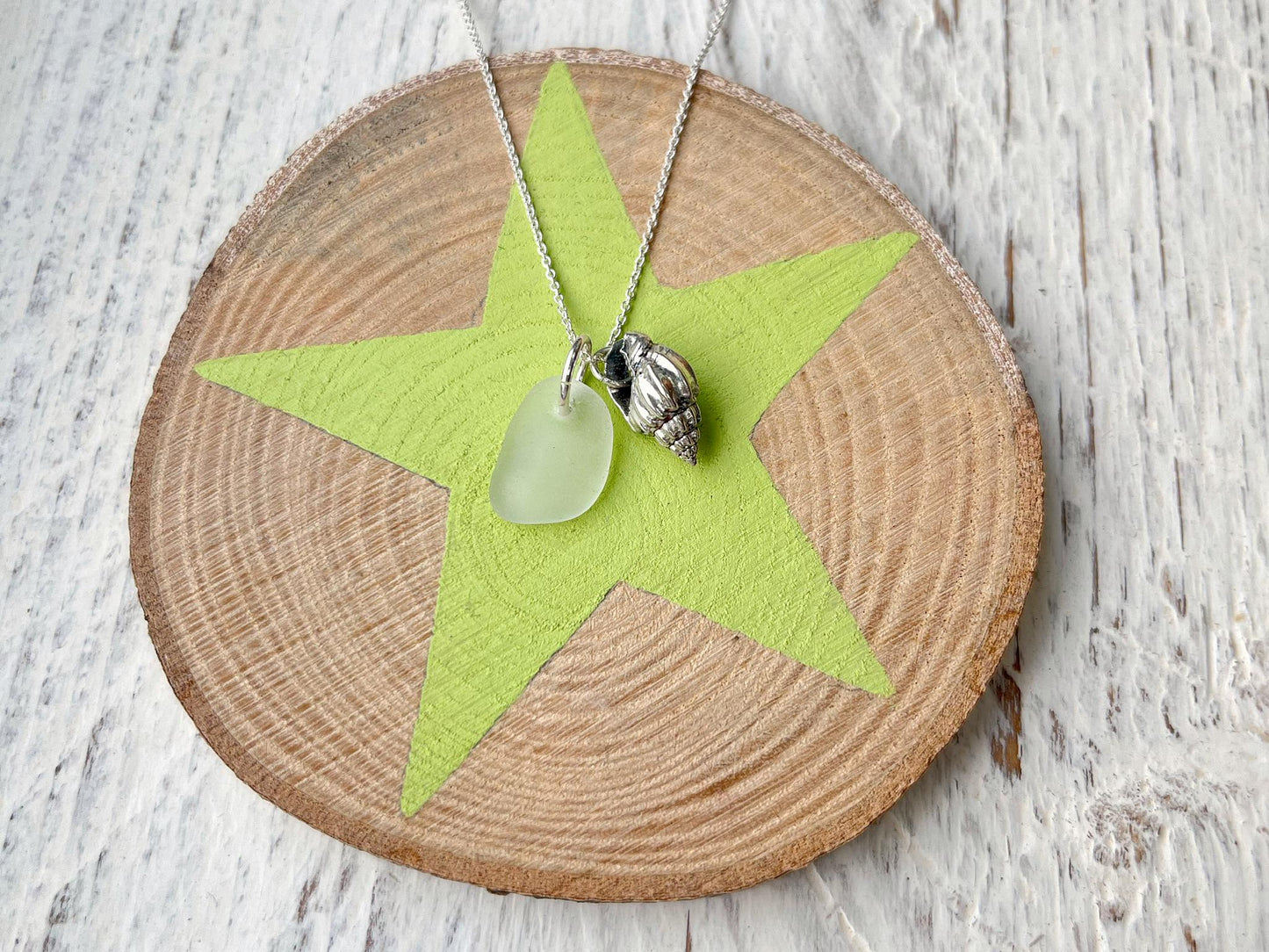 Necklace with white sea glass and silver shell pendant laid on a slice of wood with a star design against a textured surface