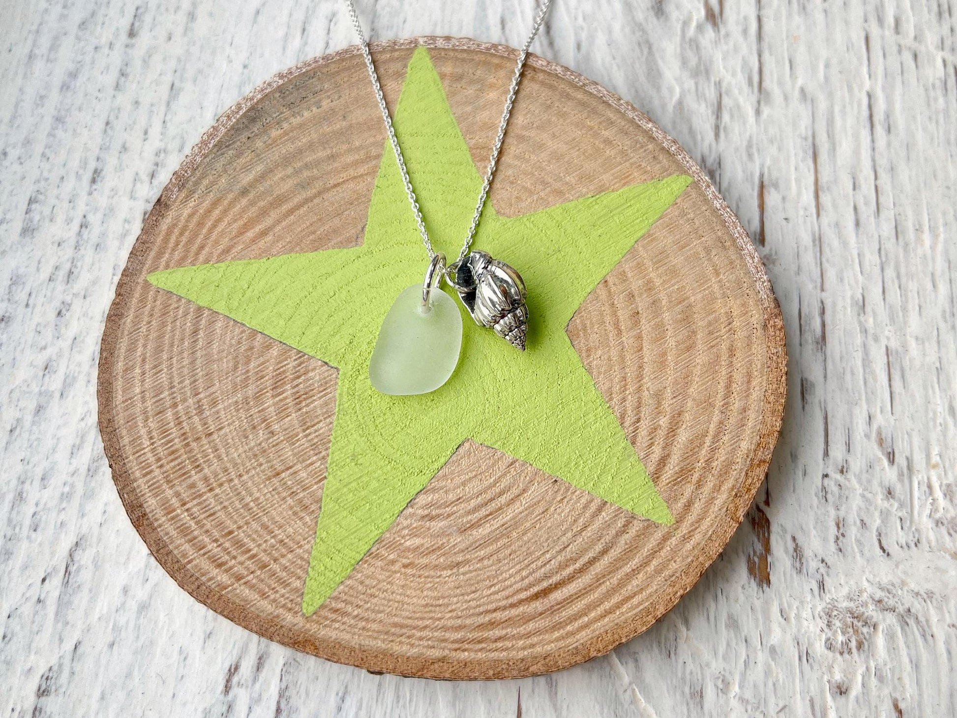 Necklace with white sea glass and silver shell pendant laid on a slice of wood with a star design against a textured surface