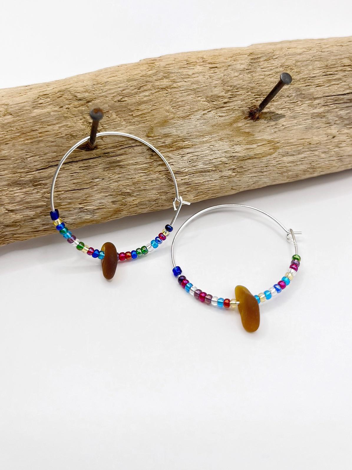 Rainbow seed bead and burnt orange sea glass hoop earrings hanging on driftwood against a white background.