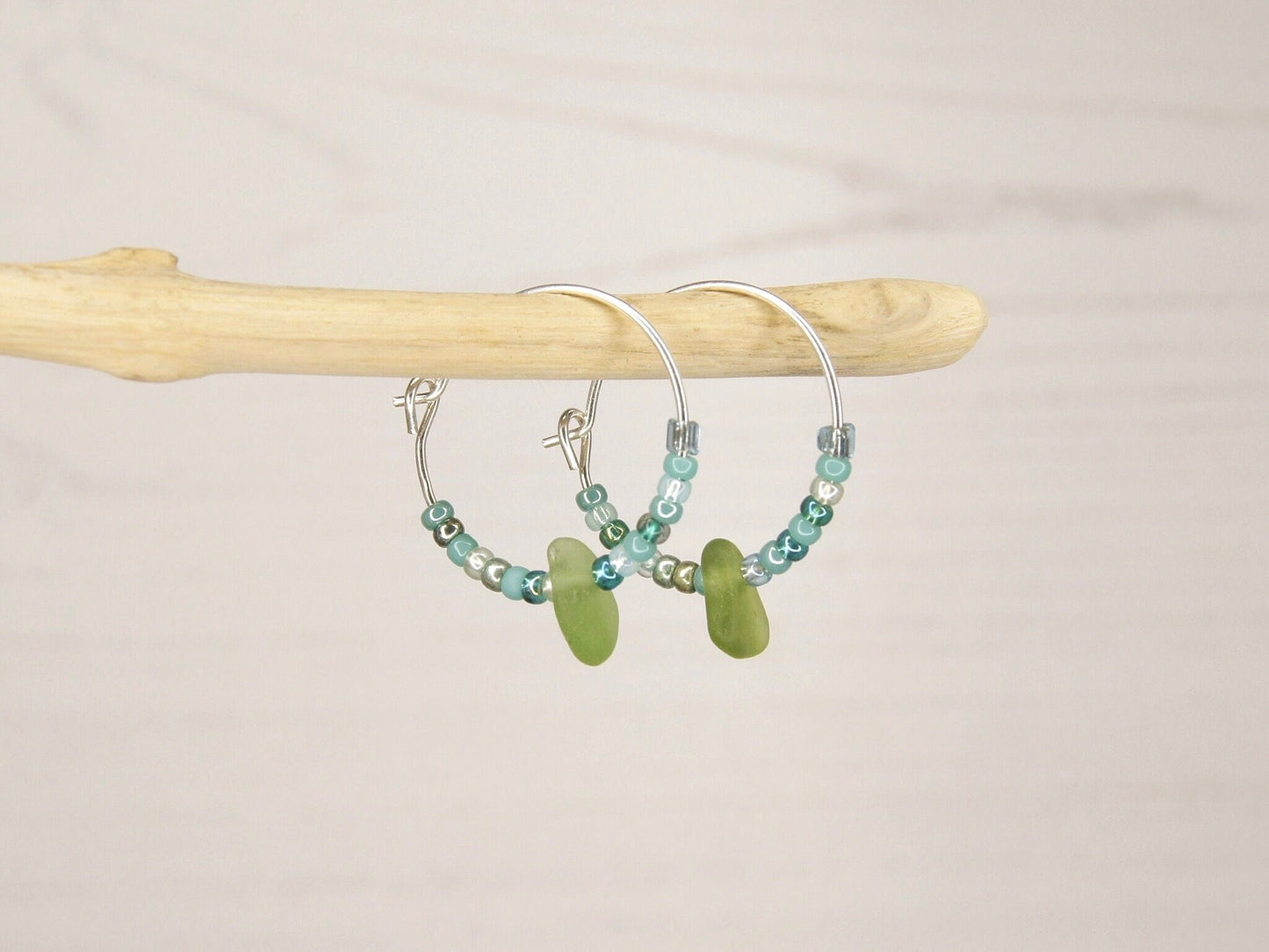 Hoop earrings with green sea glass and turquoise seed beads hanging on a driftwood against a pale background