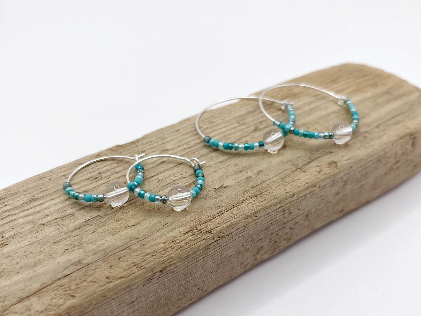 Two pairs of silver hoop earrings with turquoise seed beads and crystal laid on driftwood against a white background