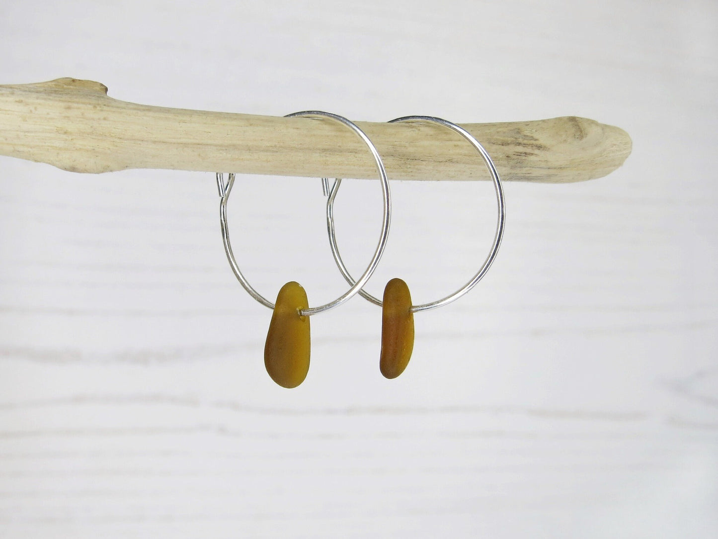 Silver hoop earrings with burnt orange sea glass hanging on a piece of driftwood against a white background
