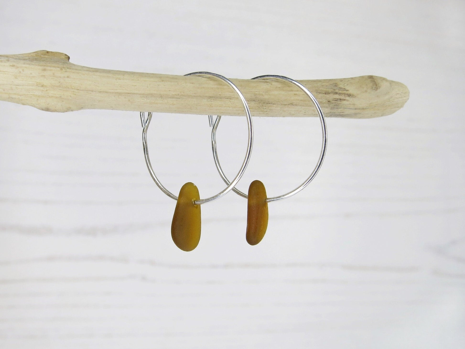 Silver hoop earrings with burnt orange sea glass hanging on a piece of driftwood against a white background