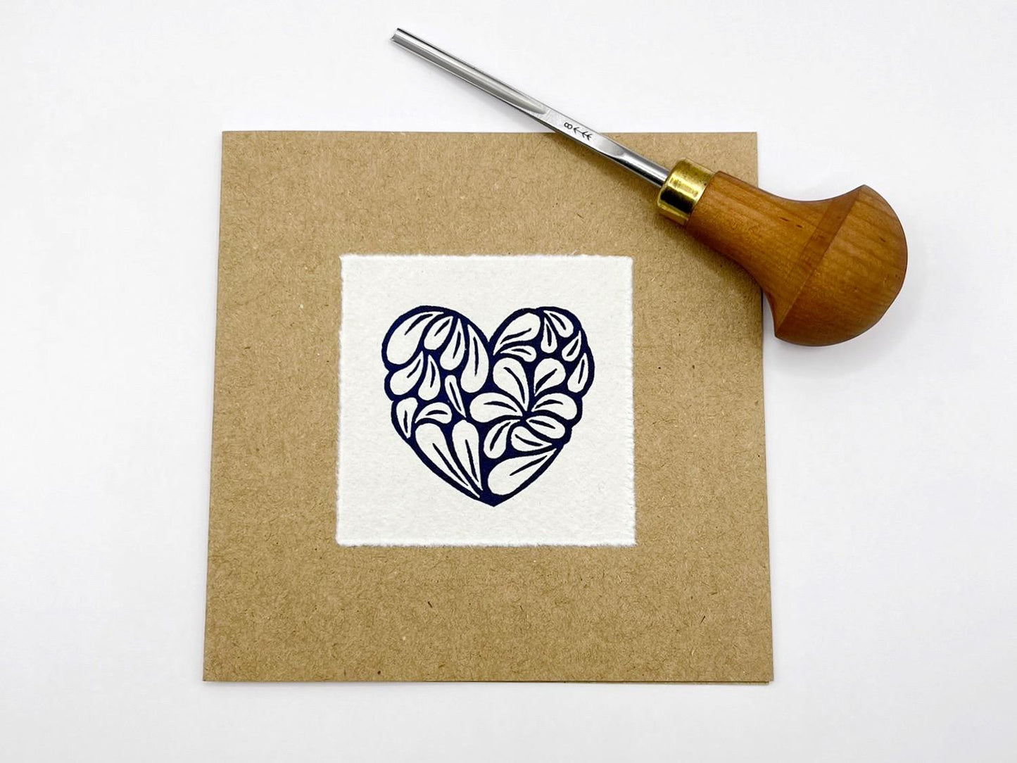 Kraft card with a white square featuring an original lino printed blue heart design, laid on a white background. Photo includes lino cutting tool.
