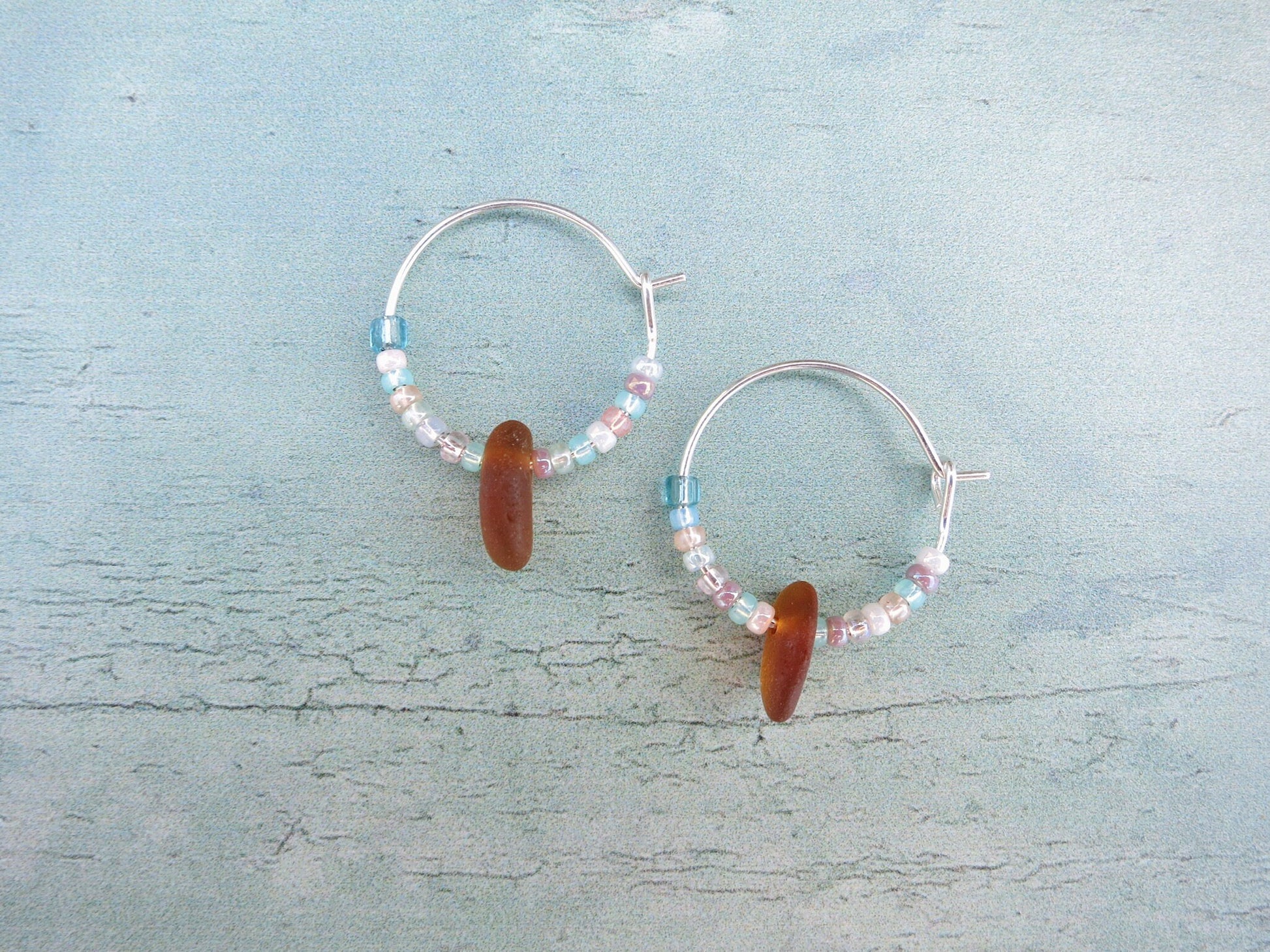 Silver hoop earrings with pastel seed beads and burnt orange sea glass laid on a light blue background